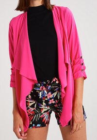 Rosa lättviktsjacka med flödande front, trekvartsärmar och rullade manschetter. Bärs över en svart topp och blommönstrade shorts.