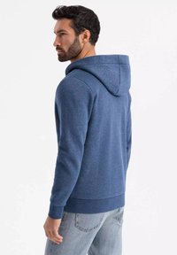 Sudadera azul marino con cordón, textura suave, puños y dobladillo acanalados. Combinada con jeans de color claro, mostrando un ajuste casual desde la parte trasera.