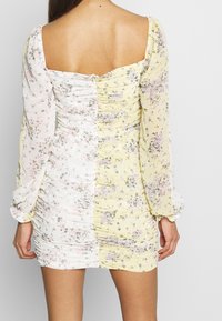 Vestido mini floral con escote cuadrado y mangas largas abullonadas. La tela es blanca y amarilla con textura fruncida, con patrones florales en rosa y morado.