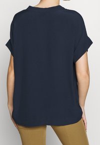 Persona che indossa una blusa corta blu scuro, svolazzante e con maniche ripiegate, abbinata a pantaloni beige, vista da dietro su uno sfondo semplice.