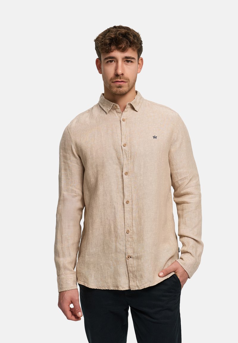 Young man with curly hair wearing a beige linen button-up shirt and black pants, standing with one hand in pocket against a plain background.