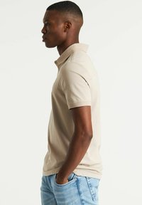 Polo beige in cotone, con colletto classico, maniche corte e finitura texture. Indossato con jeans blu.