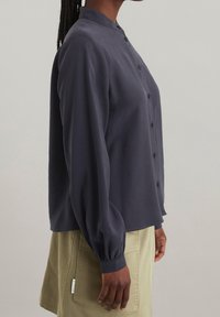 Dunkelblaues Hemd mit Mandarin-Kragen, langen Ärmeln und gerafften Bündchen, kombiniert mit khakifarbenen Shorts. Glatte Stofftextur.