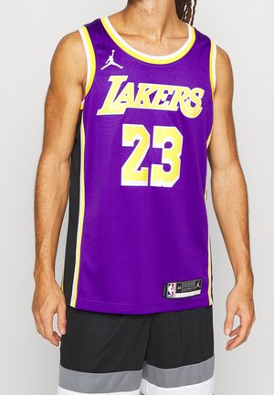 Mężczyzna w fioletowej koszulce do koszykówki Los Angeles Lakers z numerem 23 oraz czarno-białych spodenkach, stojący na jednolitym tle.