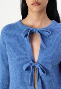 Pull en laine bleu tricoté avec une texture côtelée, doté d'un décolleté en V profond et de deux liens à l'avant au niveau du cou. Tissu doux et pelucheux.