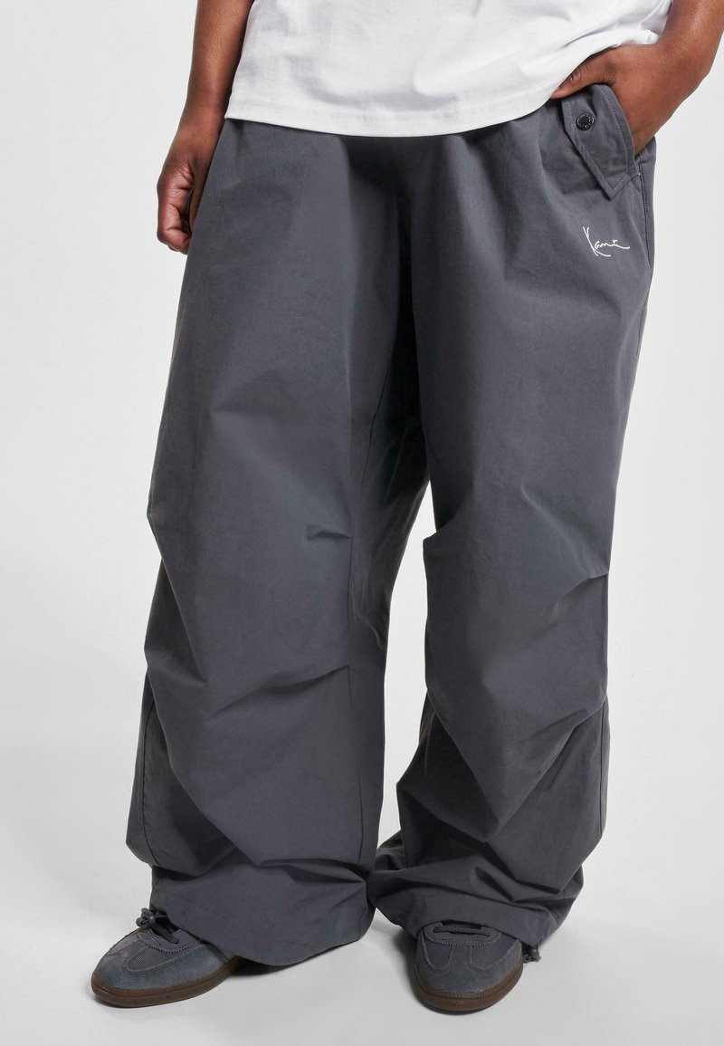 Personne portant un pantalon cargo gris foncé ample avec une poche latérale à rabat et des baskets, les mains partiellement dans les poches, un t-shirt blanc visible au-dessus de la taille.