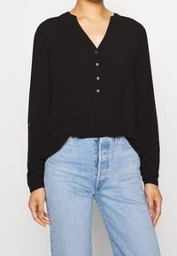 Svart långärmad blus med V-ringning, knappslå och lös passform, i kombination med högmidjade ljusblå jeans. Slät tygstruktur.