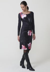 Czarna sukienka w kwiaty z długimi rękawami i węzłem w talii. Charakteryzuje się różowymi i białymi kwiatami oraz gładką teksturą materiału. Zestawiona z wysokimi na kolana czarnymi botkami.