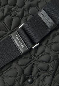 Black quilted fabric featuring heart patterns, with a black adjustable strap, textured surface, and silver hardware. Kate Spade logo visible.