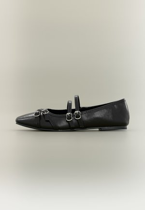 Chaussure plate noire pointue avec deux brides bouclées sur le dessus et des boucles décoratives près des orteils sur un fond neutre.