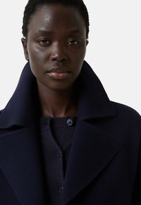 Marineblauwe wollen jas met een brede kraag, dubbelgeknoopte ontwerp en knoopaccenten, gedragen over een donker gebreide trui.