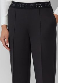 Pantalons noirs ajustés avec une taille texturée portant "BLCK" en relief avec une finition brillante. Le tissu est lisse et structuré avec des plis sur les côtés.
