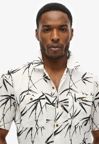 Camisa de manga corta en tela blanca con patrones de bambú negros, que cuenta con dos bolsillos en el pecho, cuello redondo y botones amarillos.