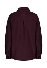 Maroon corduroy shirt with a pointed collar, buttoned cuffs, and a pleat at the back. Soft texture and oversized fit, accentuating the design.