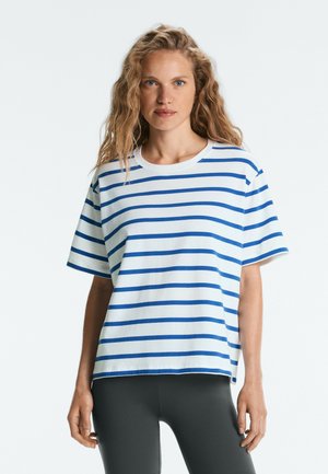Femme aux longs cheveux bouclés portant un t-shirt blanc à manches courtes avec des rayures horizontales bleues et des leggings foncés sur un fond uni.