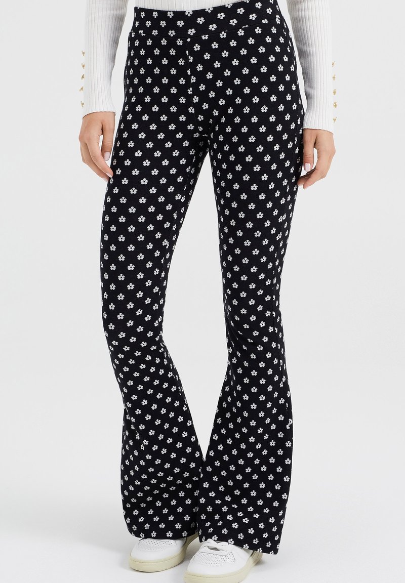 Černé rozšířené kalhoty s bílými květinovými vzory, vyrobené z měkkého materiálu. Pas je elastický a kalhoty jsou v kombinaci s bílými teniskami.