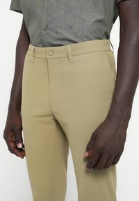 Pantalones de tela caqui clara con una textura suave. Cuentan con un bolsillo frontal y una cinturilla con cierre de botón. Combinados con una camisa gris con estampado.