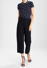 Polo shirt blu navy con bordi bianchi e strisce diagonali, abbinato a pantaloni neri a vita alta e a gamba larga e sandali neri con cinturini.