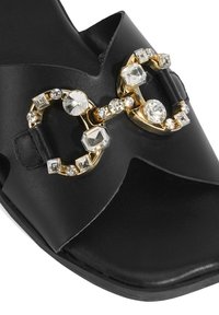 Sandale glissante en cuir noir ornée d'un embellissement accentué d'or, avec des pierres transparentes et texturées disposées en un motif décoratif.