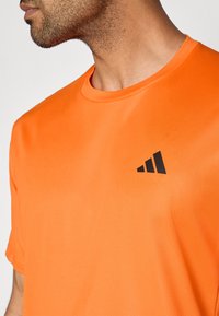 Camiseta deportiva de color naranja brillante con un acabado texturizado. Presenta un cuello redondo y un logo negro en el pecho izquierdo. Mangas cortas.