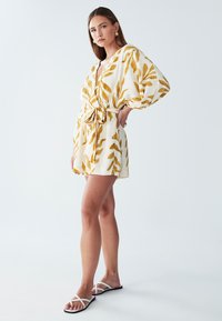 Wit jurkje met korte mouwen en gele bladpatronen, getailleerd bij de taille. Draagt witte sandalen met bandjes, tegen een lichte achtergrond.