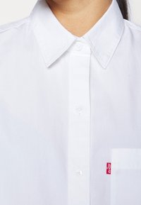 Gros plan d’une chemise blanche à col boutonnée, avec une poche poitrine et une étiquette rouge avec le logo Levi’s sur la poche.
