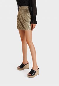 Mini gonna d'oro metallico con taglio dritto. Sandali con tacco a cuneo neri, con piattaforma in paglia intrecciata e design aperto sulla punta. Blusa nera trasparente con maniche lunghe.