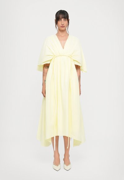 Gelbes, fließendes Kleid mit tiefem V-Ausschnitt, geraffter Taille und drapierten Ärmeln. Mit einem hohen tiefen Saum und leichtem Stoff. Getragen mit weißen Absätzen.