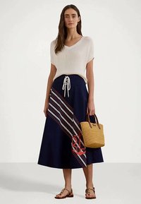 Γυναίκα που φοράει λευκό κοντομάνικο πλεκτό τοπ, ναυτικό midi φούστα με διαγώνιες ρίγες και σχέδιο άγκυρας, κρατώντας τσαντάκι από πλεγμένο υλικό, φορώντας πέδιλα.