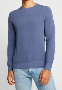 Blå stickad tröja med rund halsringning, strukturerat mönster, ribbad mudd och nederkant. Har en liten logotyp på bröstet.