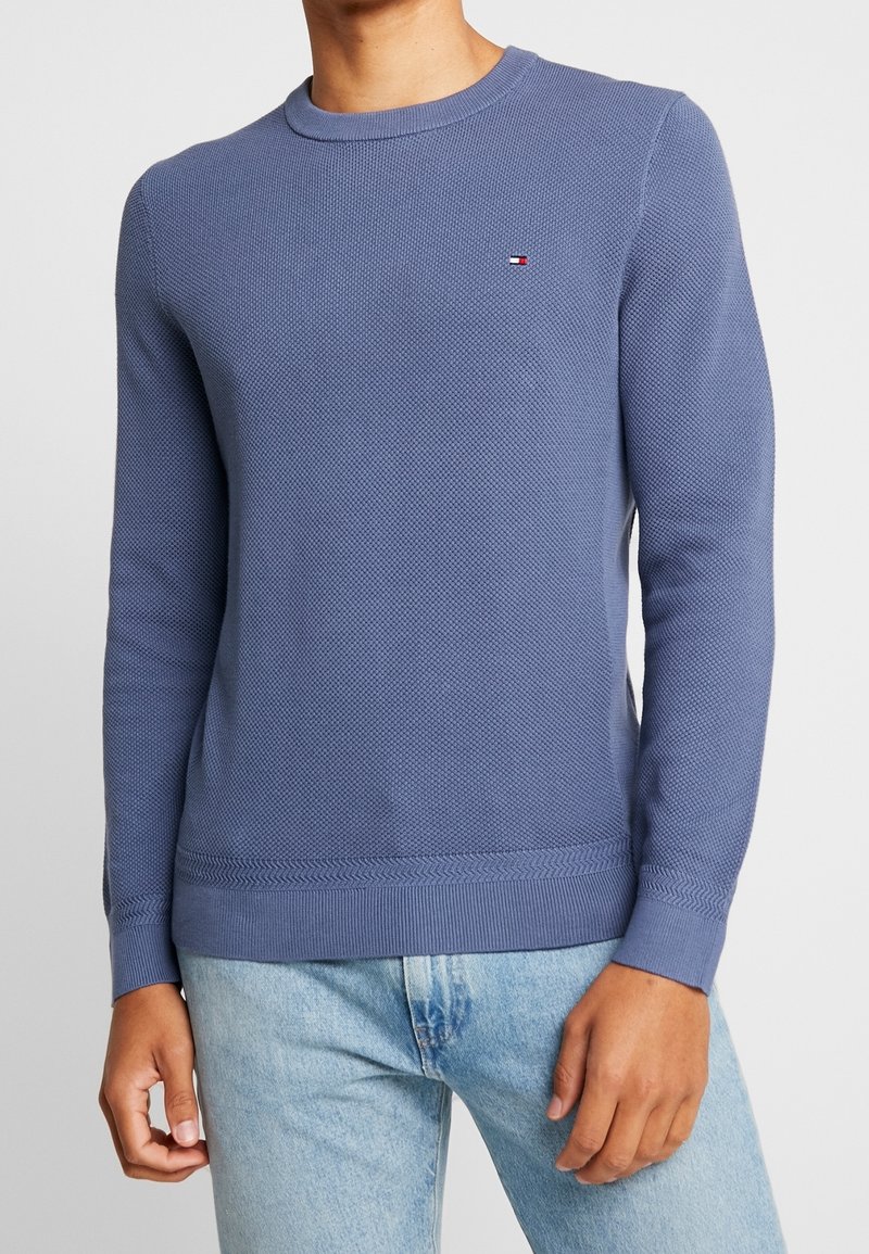 Blå stickad tröja med rund halsringning, strukturerat mönster, ribbad mudd och nederkant. Har en liten logotyp på bröstet.