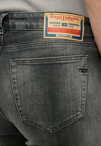 Pogled s leđa na osobu koja nosi izblijedjele crne Diesel traperice s pravokutnim kožnim oznakom marke iznad desnog stražnjeg džepa.