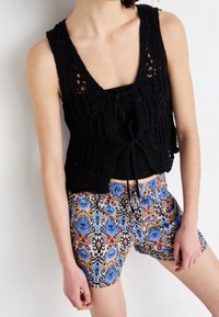 Gilet nero all'uncinetto con tessuto a trama aperta, abbinato a pantaloncini multicolori con motivi e dettagli blu e oro.