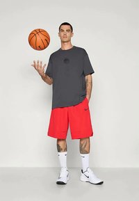 Camiseta gris de manga corta con un logo redondeado, combinada con pantalones cortos rojos sueltos, calcetines blancos y zapatos deportivos negros. Sosteniendo un balón de baloncesto.