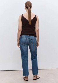 Mujer de espaldas a la cámara, vestida con una blusa negra sin mangas, jeans azules, sandalias negras y peinado en una coleta.