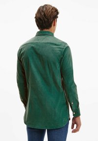 Camisa de manga larga en algodón verde oscuro con cuello abotonado, costuras visibles y puños con botones, con un dobladillo curvado.