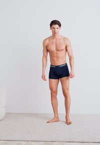 Marinefarbene Boxershorts aus dehnbarem Material, mit einem kontrastierenden Bund mit "Lacoste"-Logo. Das Modell steht vor einem neutralen Hintergrund.