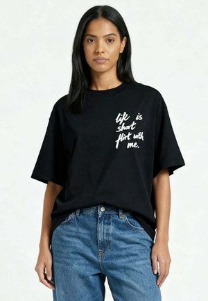 Donna con capelli neri lisci indossa una T-shirt nera oversize con la scritta bianca "la vita è breve, flirtami" e jeans blu.