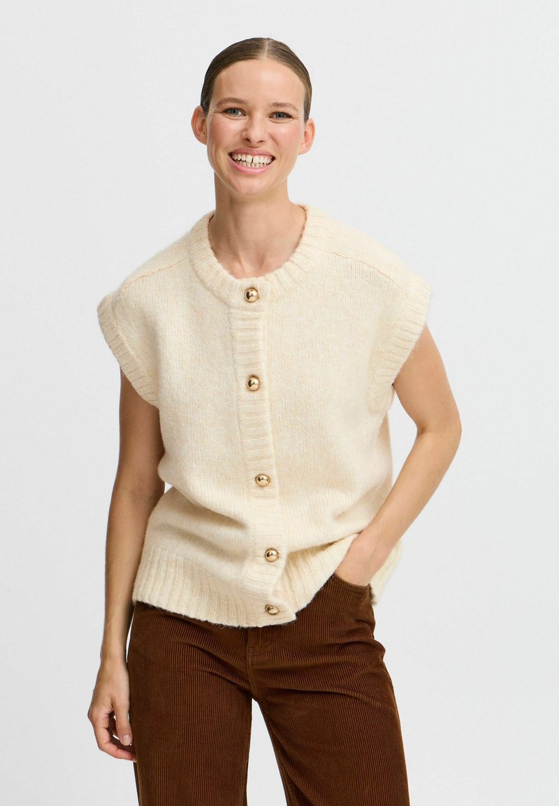 Beige gestrickte Knopfveste mit kurzen Ärmeln, featuring einem gerippten Saum und großen glänzenden Knöpfen, kombiniert mit braunen Cordhosen.