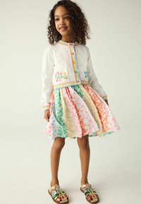 Maglione bianco all'uncinetto con bottoni colorati e ricami floreali, abbinato a un vestito a campana floreale multicolore e sandali metallici.