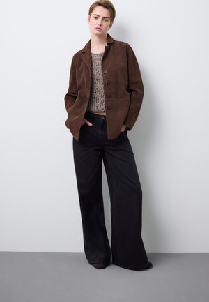 Bruine suède jas met een reverskraag en knoopsluiting, gedragen over een gestructureerde gebreide trui en losse zwarte broek met wijde pijpen.