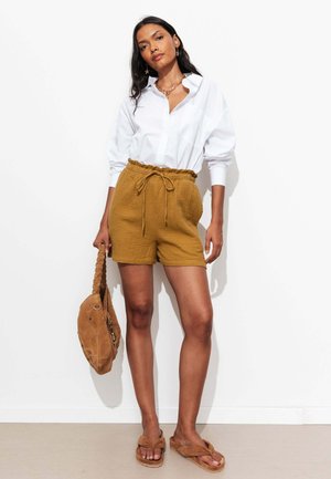 Femme portant une chemise blanche à manches longues, un short moutarde à cordon, des sandales beige, tenant un sac à main beige avec une anse tressée, debout contre un mur blanc.