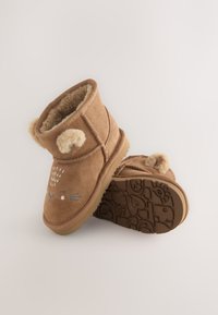 Bottes cheville en daim marron avec accents d'oreilles duveteuses, traits du visage illustrés, et semelles en caoutchouc texturées avec des motifs ludiques.