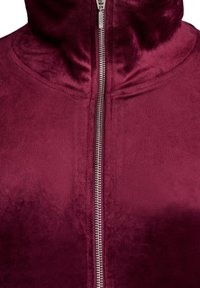 Burgundska zip-jaka iz mehkega, plišastega materiala. Ima visok ovratnik in gladek srebrn zadrgo. Teksturna površina z rahlim leskom.