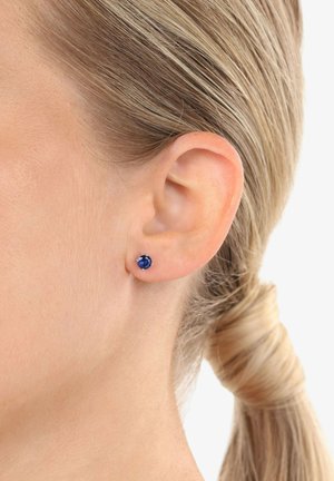 Pendientes de oro con una gema azul redonda, montados en un diseño simple de cuatro garras, mostrados en la oreja de un modelo.