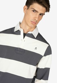 Polo a maniche lunghe a righe in grigio e bianco. Presenta un colletto bianco, patta con bottoni e un piccolo logo sul petto. Materiale in cotone.