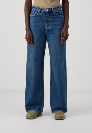 Bredde ben jeans i blått, med høy midje, fem lommer og subtil falming. Kombinert med olivengrønne sko.