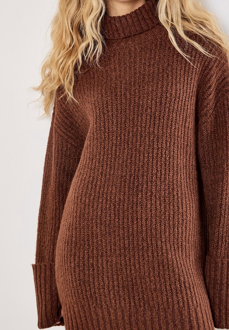 Personne blonde portant un pull en maille côtelée épaisse marron avec un col montant et des manches longues.