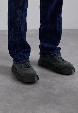 Person trägt dunkelblaue Jeans und schwarze Turnschuhe und steht auf grauem Boden vor neutralem Hintergrund.