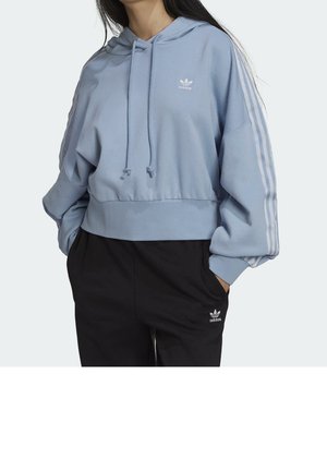 Frau trägt hellblauen Adidas Cropped-Hoodie mit drei weißen Streifen an den Ärmeln und schwarze Adidas Jogginghose mit Logo.
